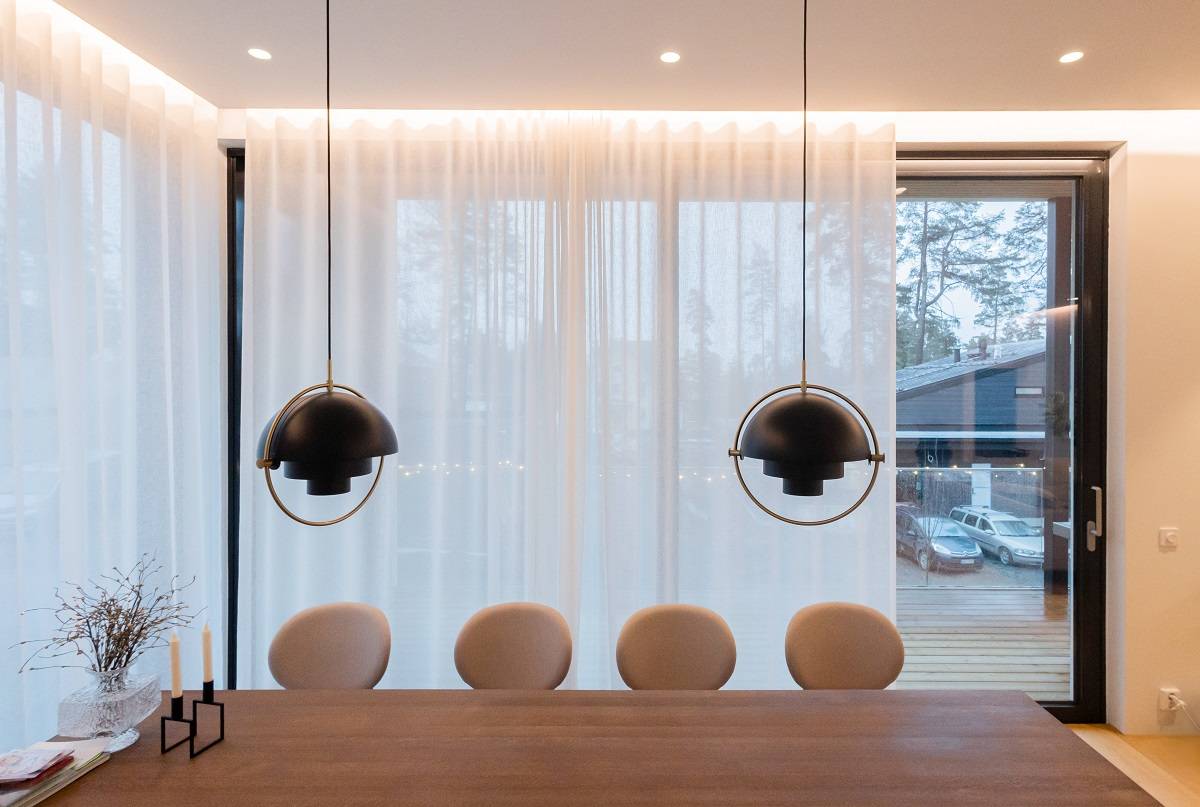 El comedor está iluminado con tiras de LED detrás de cortinas de luz natural, focos empotrados en el techo y dos lámparas colgantes sobre la mesa.